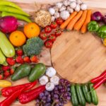 A variety of fresh fruits and vegetables arranged around a round wooden cutting board